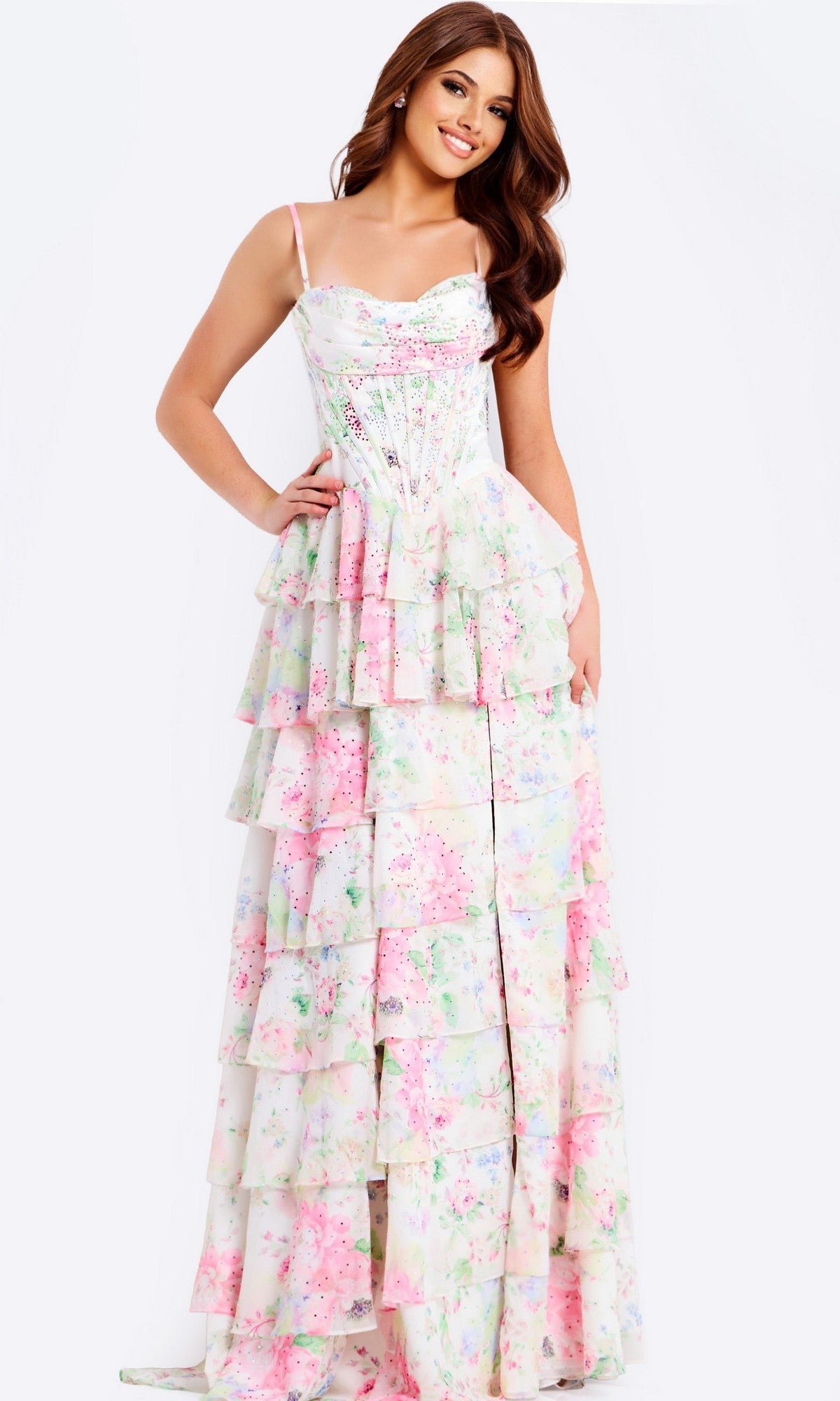 A woman stands and smiles against a plain white background wearing the Jovani 47282 long prom dress featuring pastel floral prints, a corset bodice, and tiered ruffles.